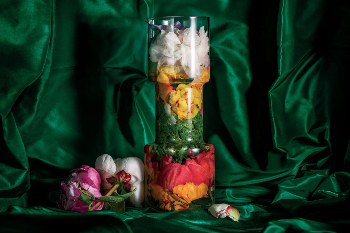 A glass decanter filled with layers of marigold, hibiscus, linden, cherry blossom and peony, which is also scattered on the green taffeta. Photography by Anthony Cotsifas.